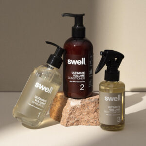 A pack shot of Swell volume shampoo, conditioner, and root complex on a light stone background