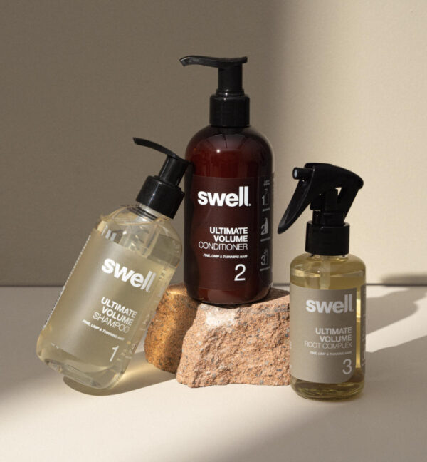 A pack shot of Swell volume shampoo, conditioner, and root complex on a light stone background